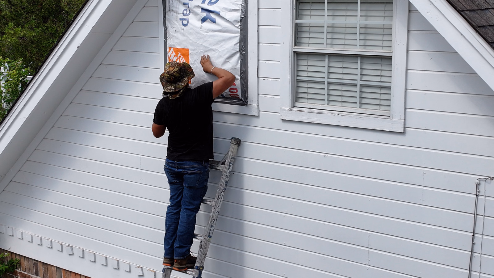 Worker on a ladder installing house wrap around a window opening during a window replacement.