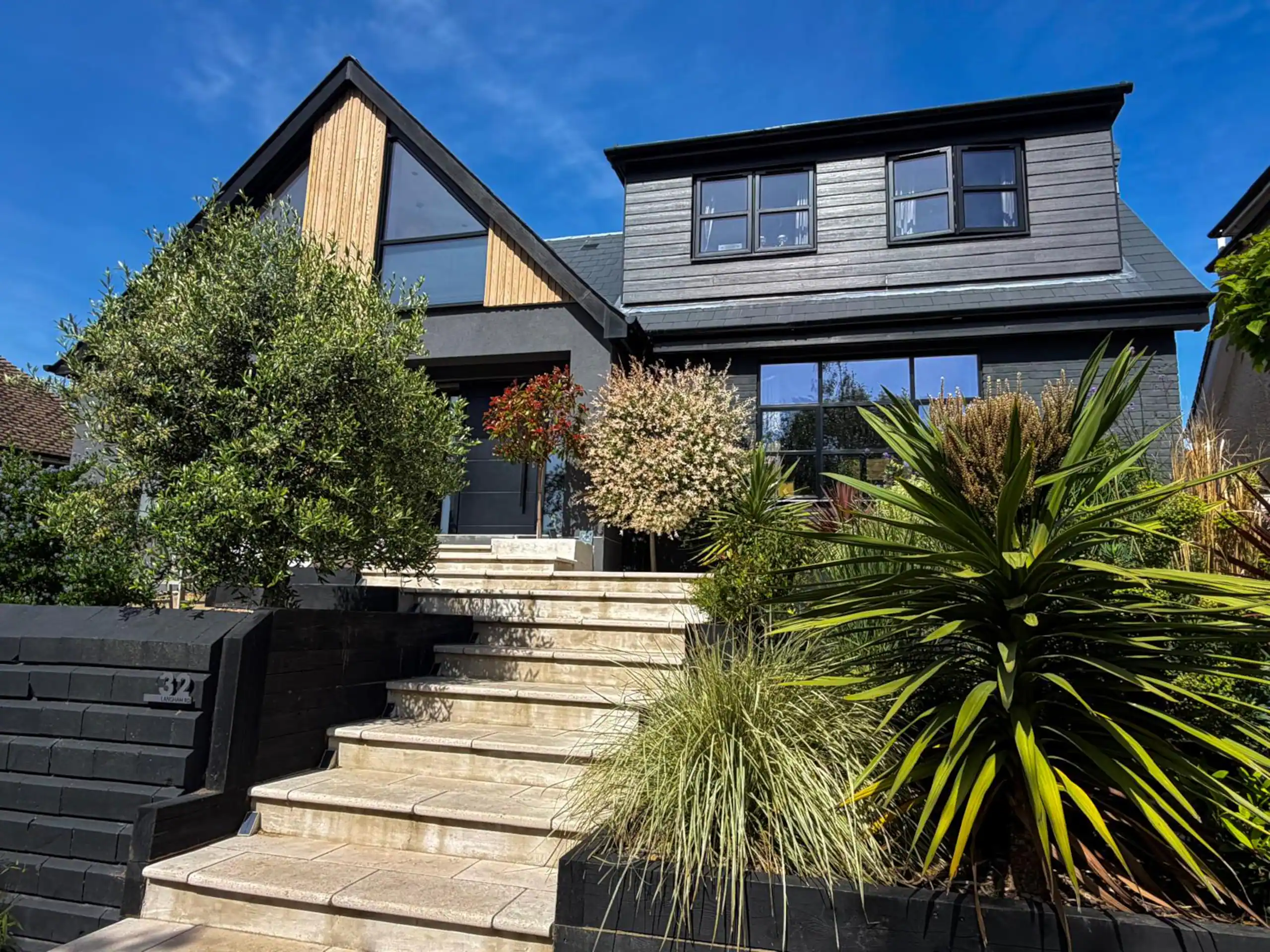 Modern two-story house with black siding, large windows, stone steps, and lush greenery.