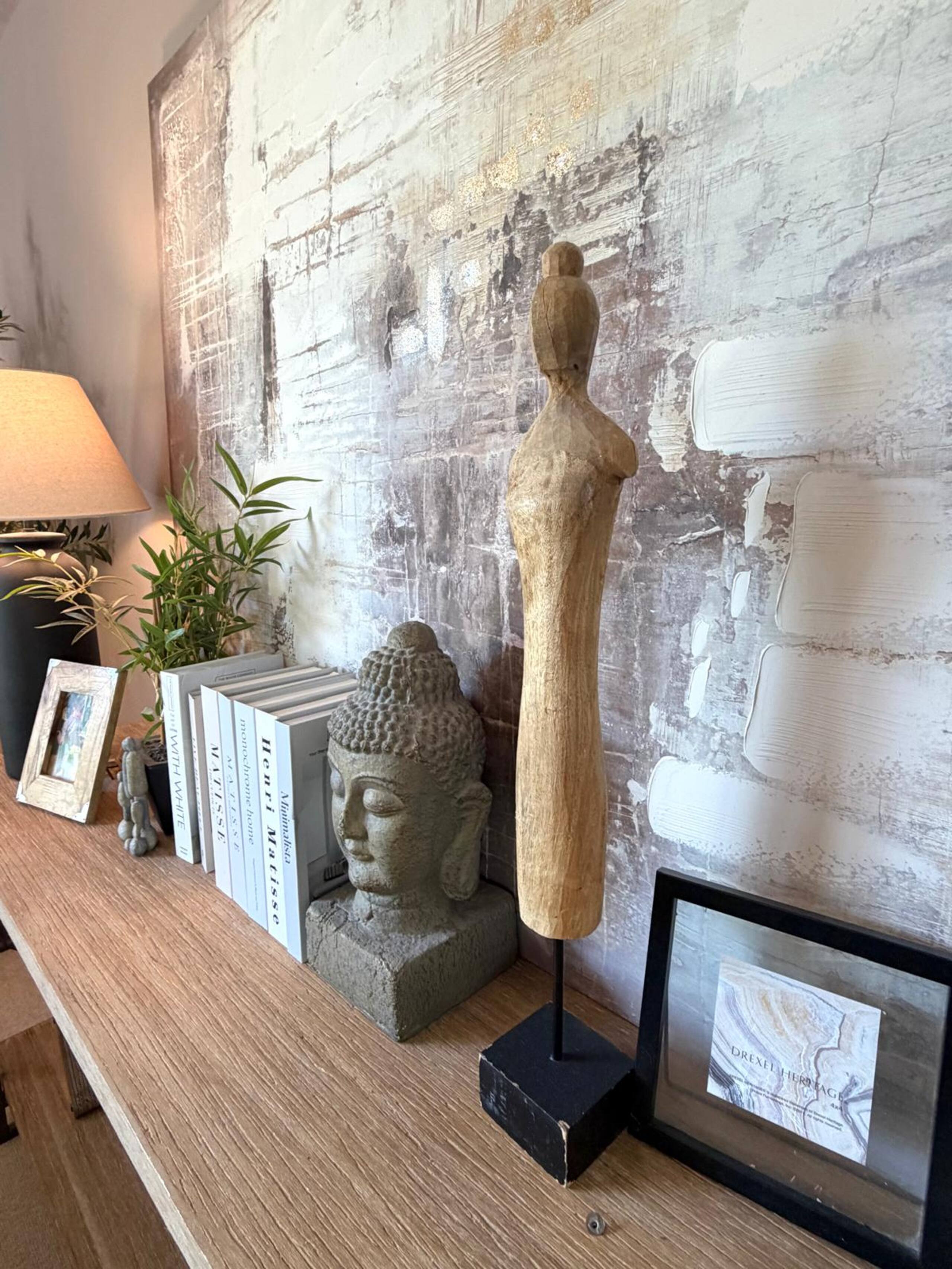 Wooden table with Buddha head statue, wooden figure, books, a lamp, plant, and framed artwork.