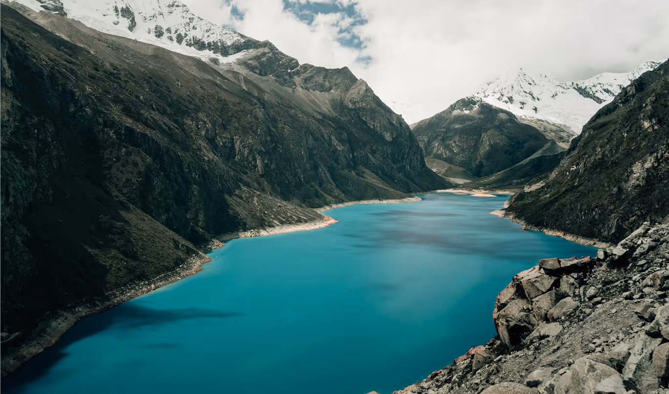 Turquoise lake surrounded by rocky mountains with snow-capped peaks under a cloudy sky.