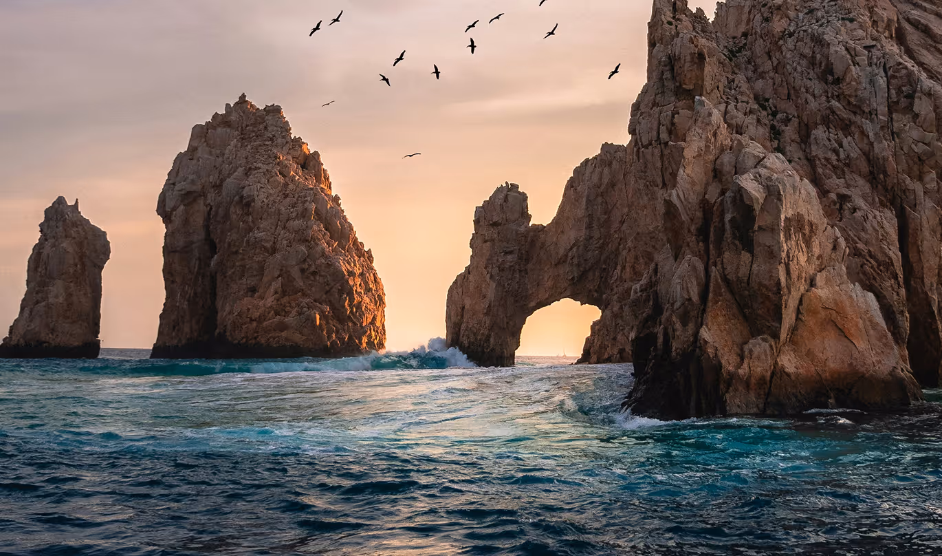 Rock formations in the ocean with a natural arch and birds flying at sunset.