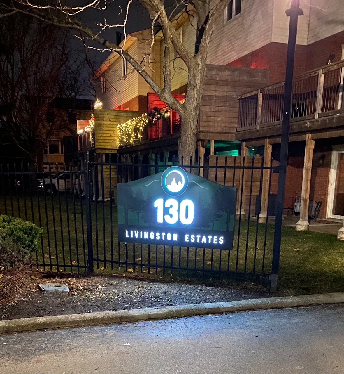 Illuminated sign on black fence reads 130 Livingston Estates in front of a house at night.