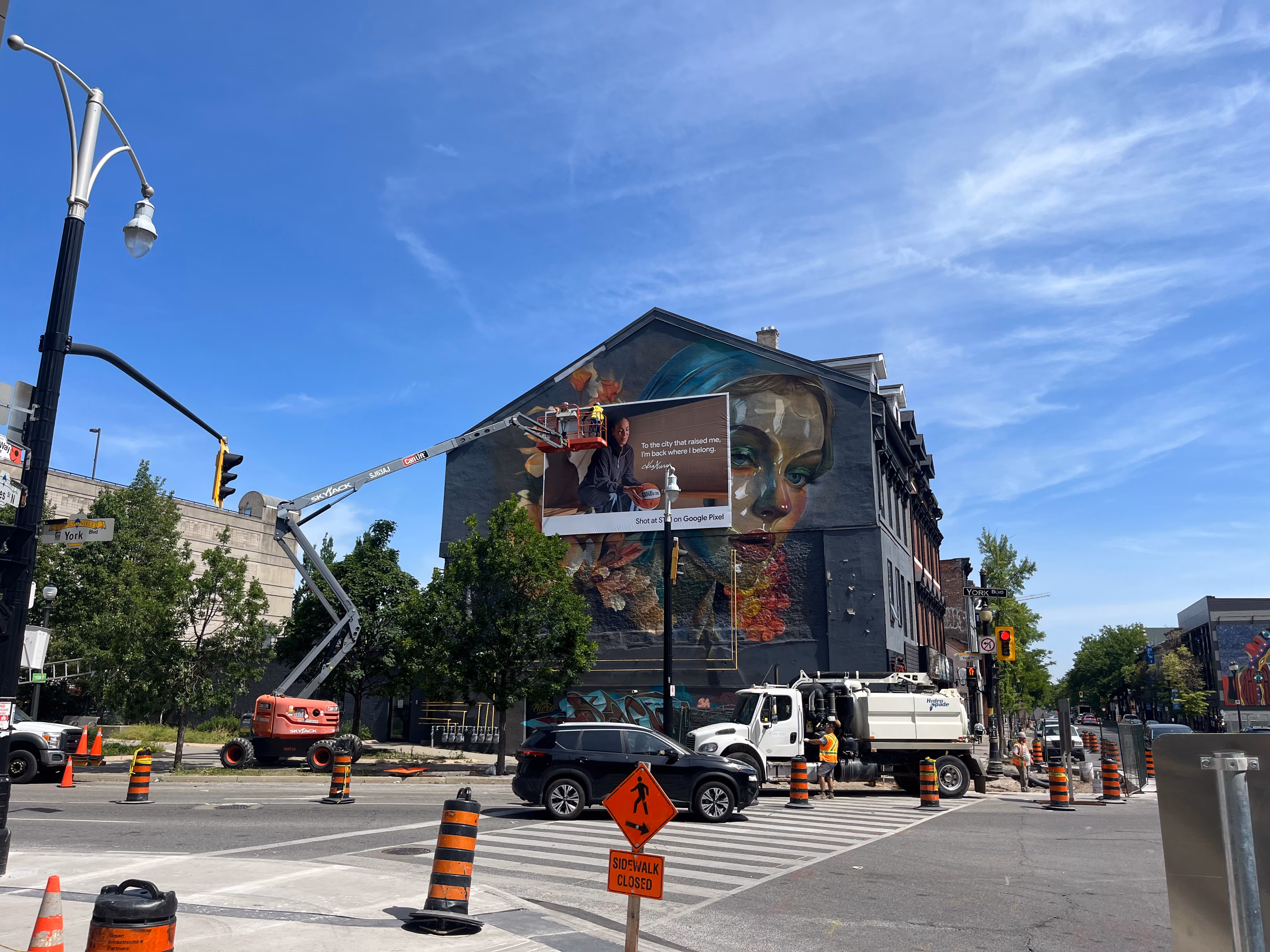 Workers on lift installing a billboard on a building with a large mural, street closed for construction.