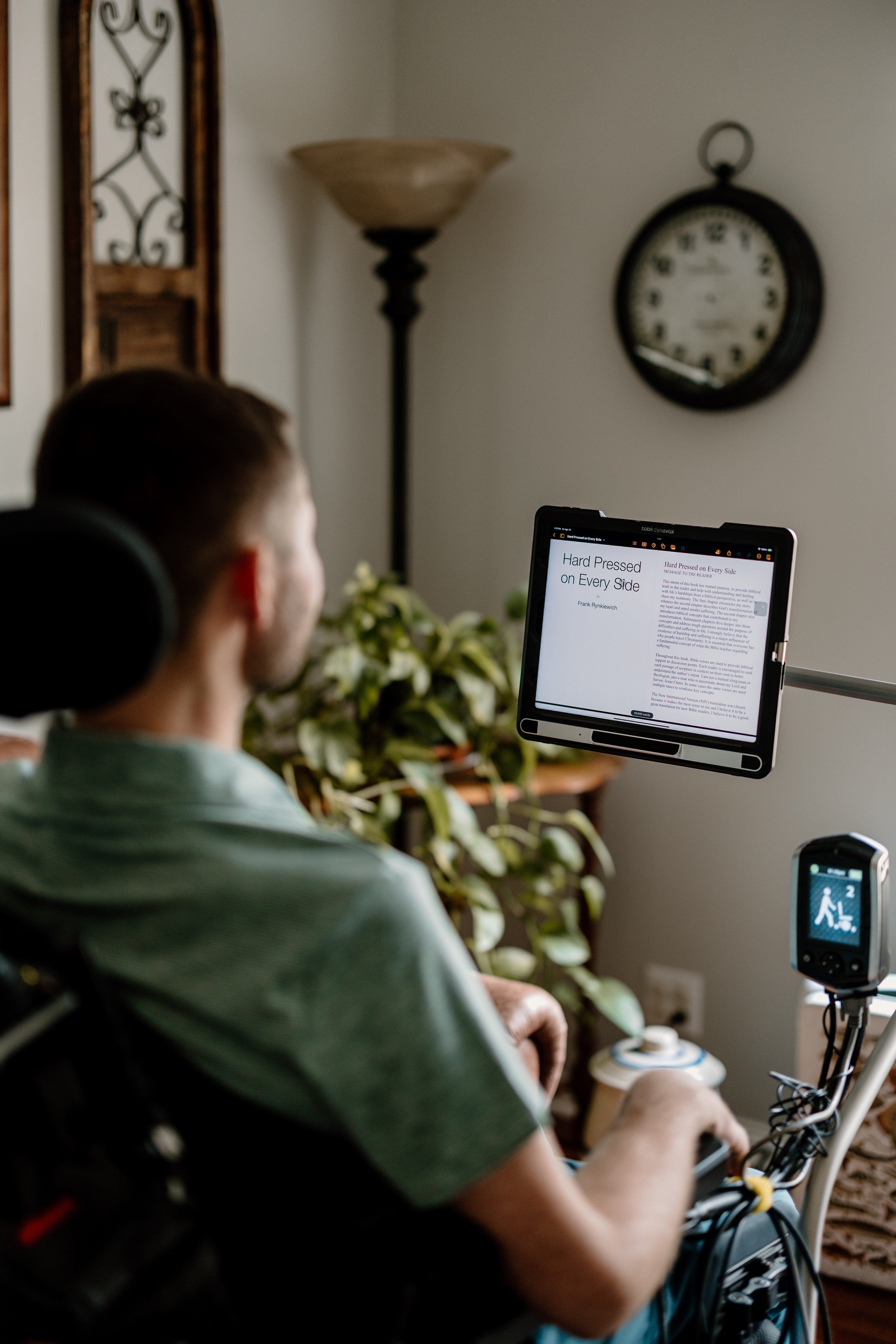 Man in wheelchair reading text on mounted screen with a clock and plants in the background.