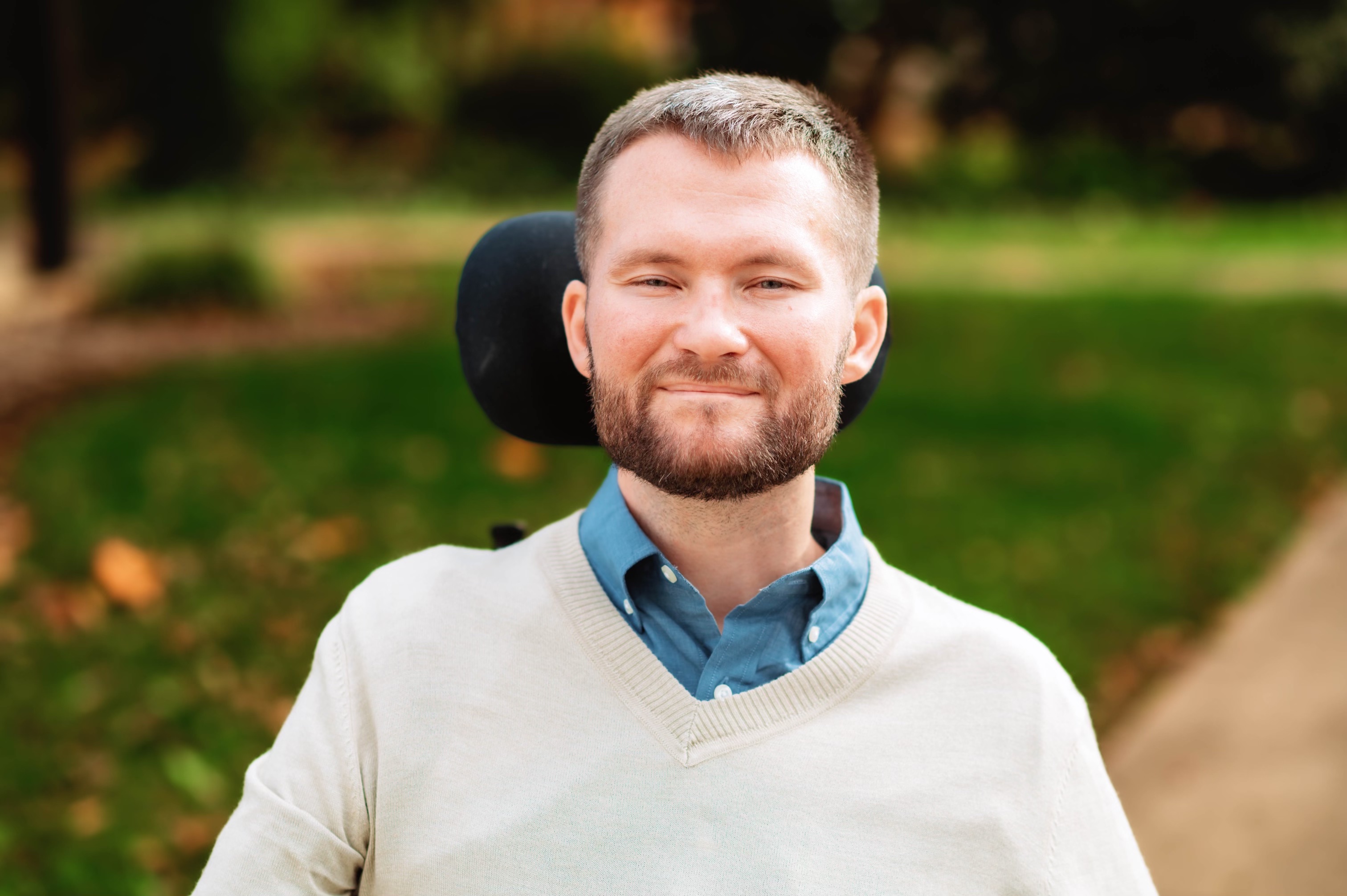 Bearded man smiling outdoors wearing a beige sweater and blue collared shirt, seated in a wheelchair.