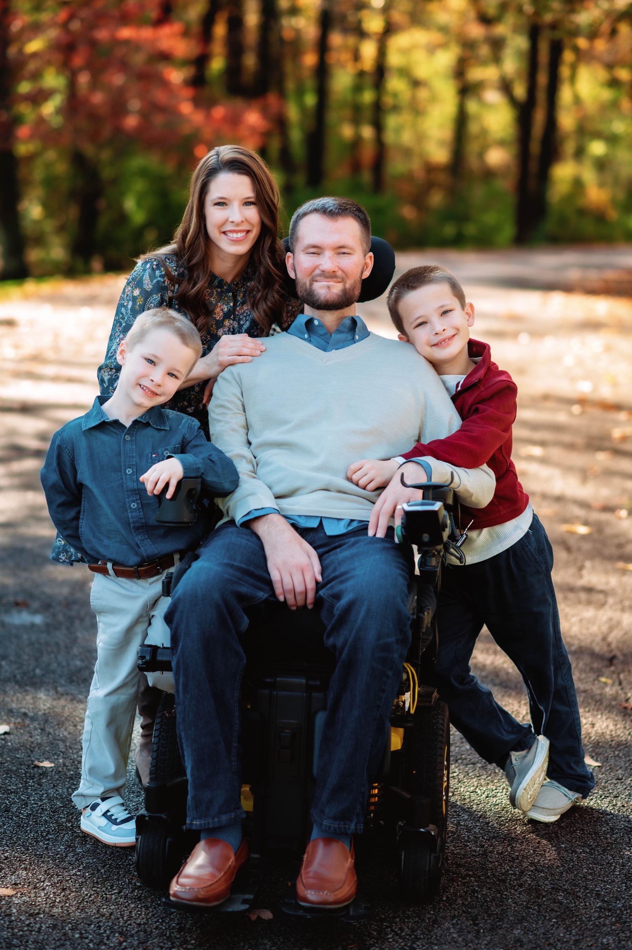 Smiling family of four with father in wheelchair outdoors on fall day with colorful trees.