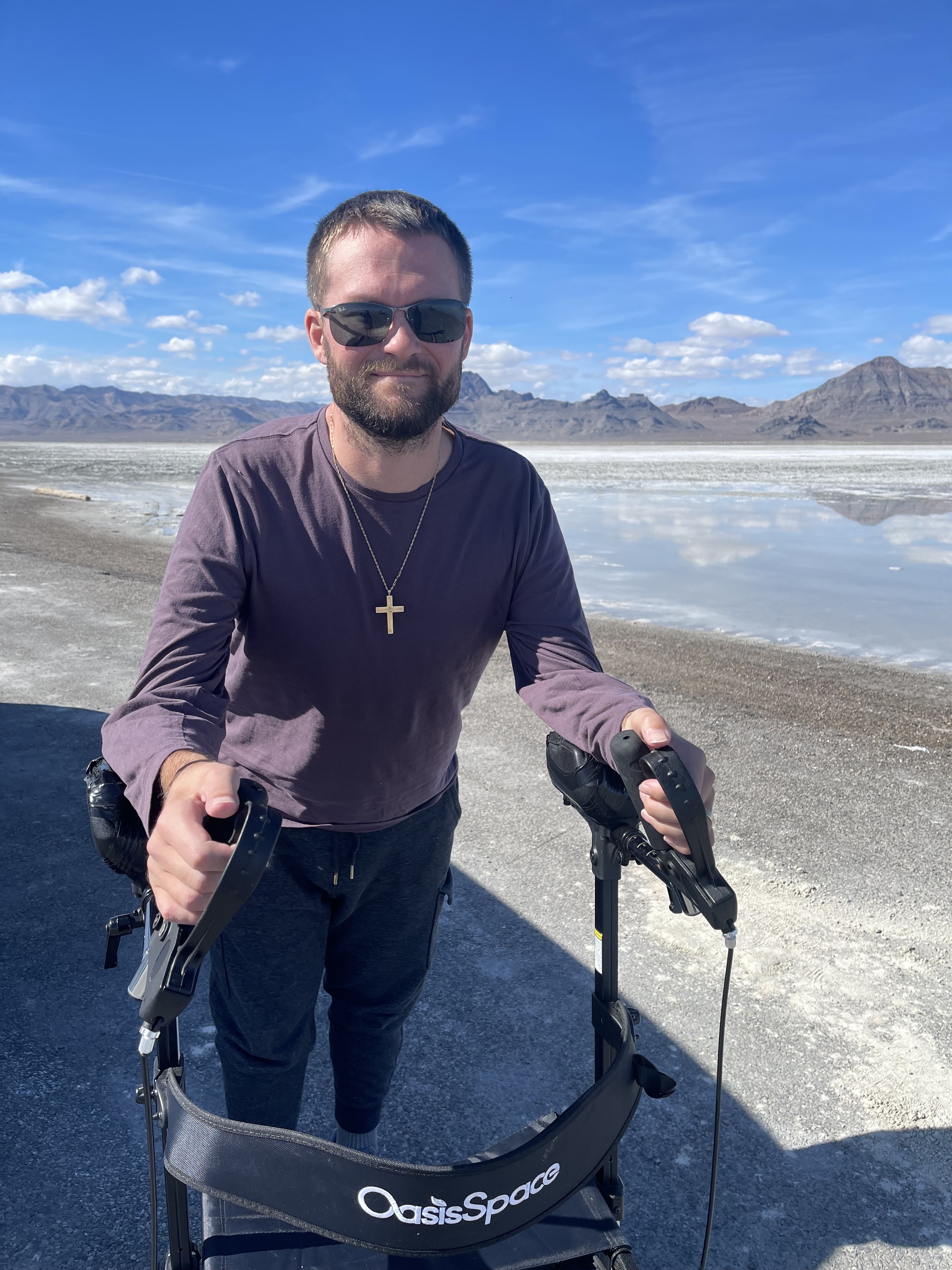 Man wearing sunglasses and a cross necklace outdoors using an OasisSpace walker near a salt flat.