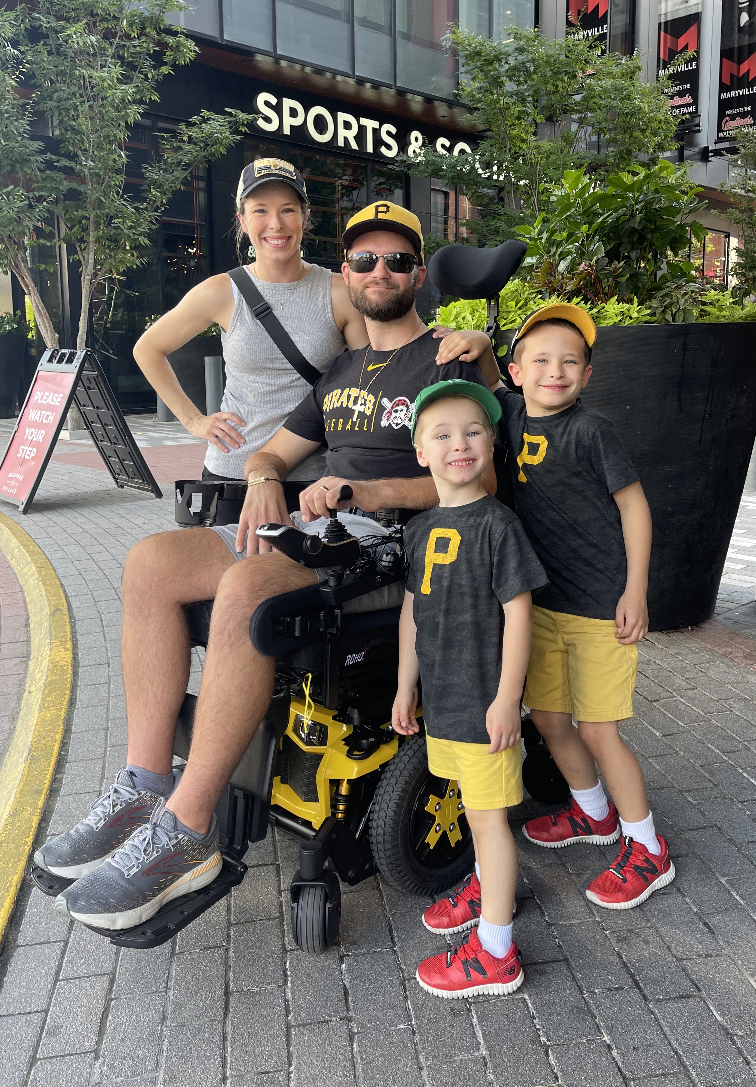 Family of four outside sports store, man in wheelchair wearing Pirates gear, two boys in matching outfits.