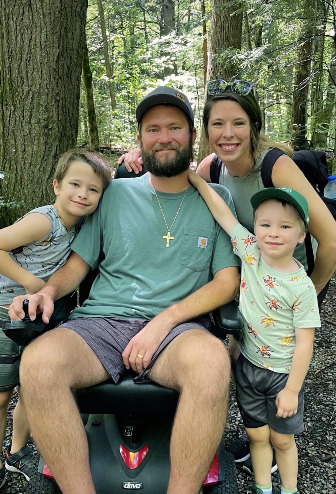 Happy family of four posing in a forest, father seated on a mobility scooter with kids and mother hugging him.