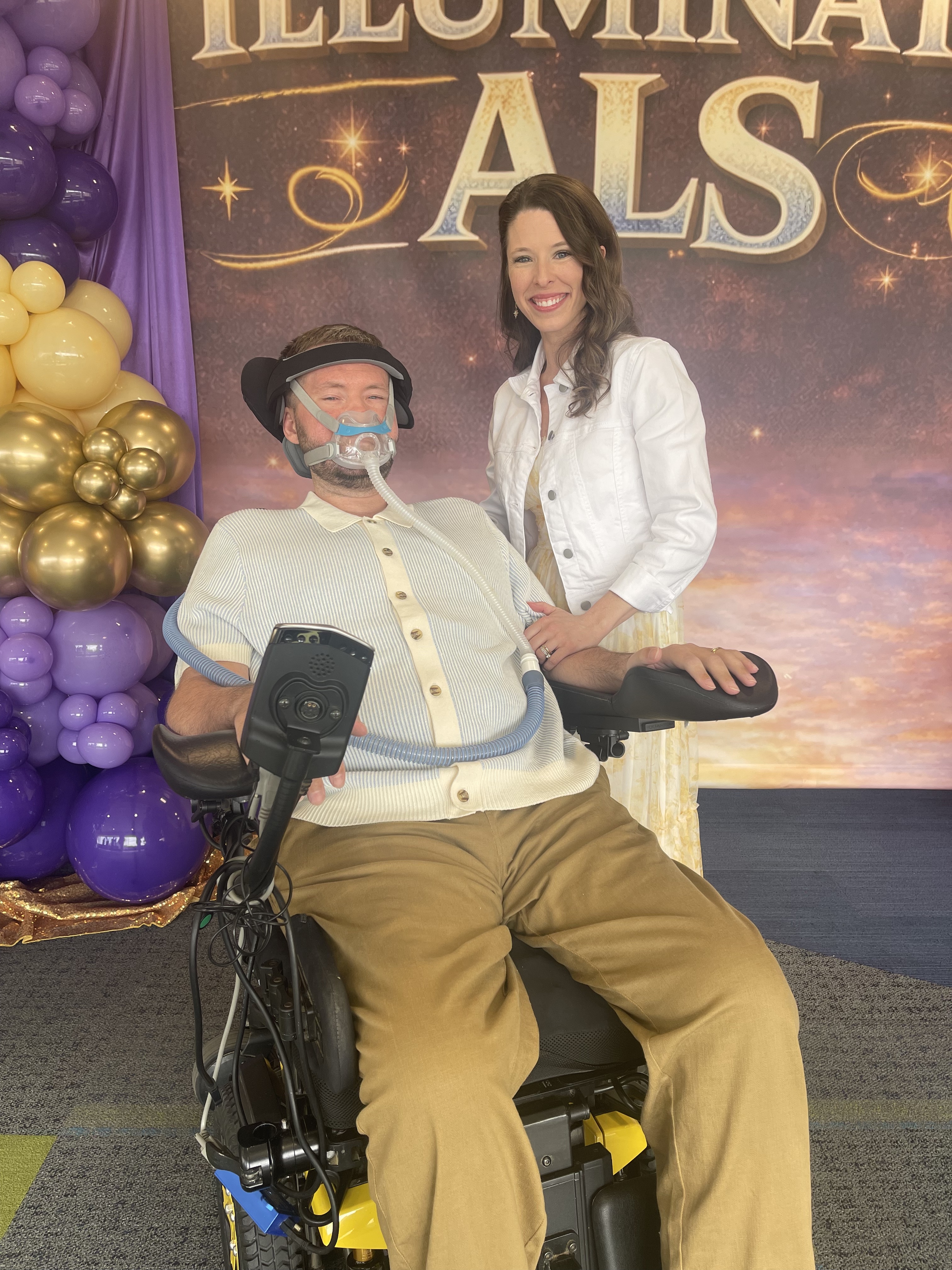 Man in wheelchair with breathing mask and woman in white jacket beside him at ALS event.
