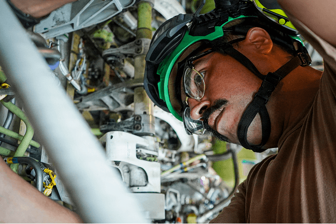 Young technician in a green helmet and safety glasses inspecting aircraft engine components up close