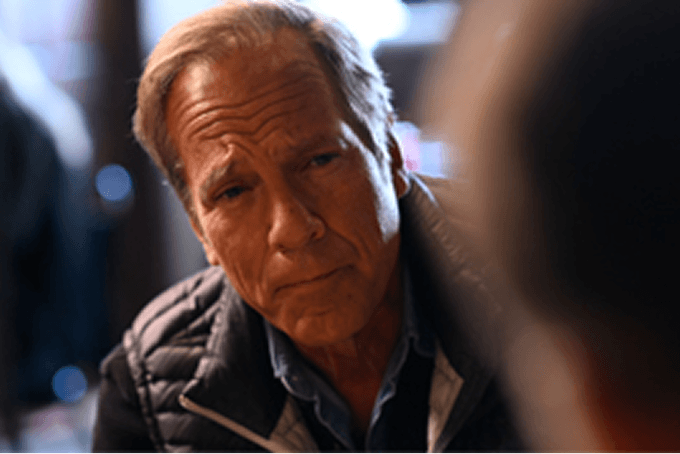 Mike Rowe in a dark jacket listening intently during a conversation