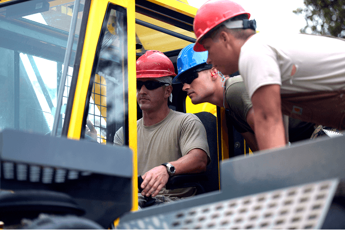 Three apprentices in hard hats and military uniforms training on heavy construction equipment