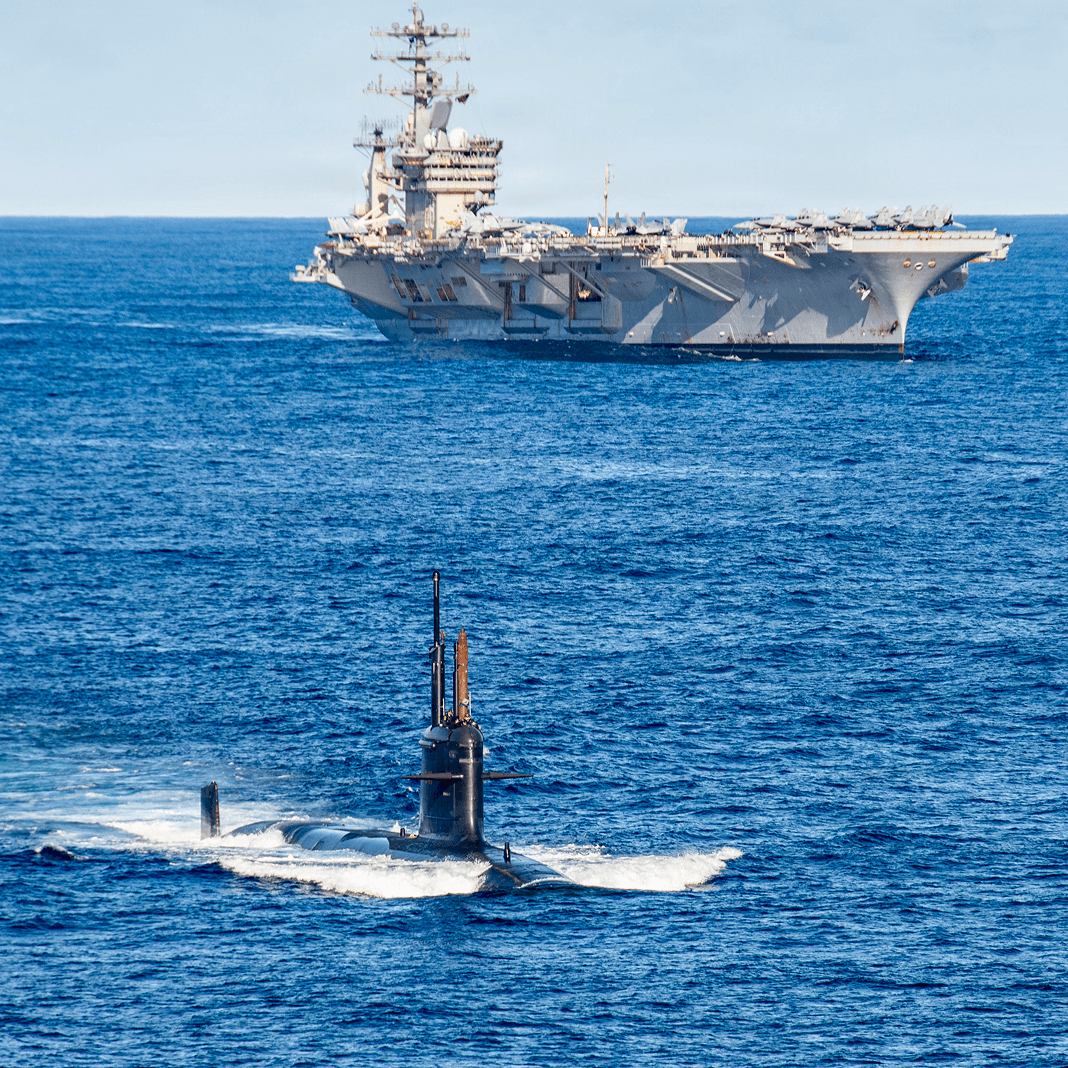 A submarine moves through open ocean on the surface, with a large aircraft carrier in the background.