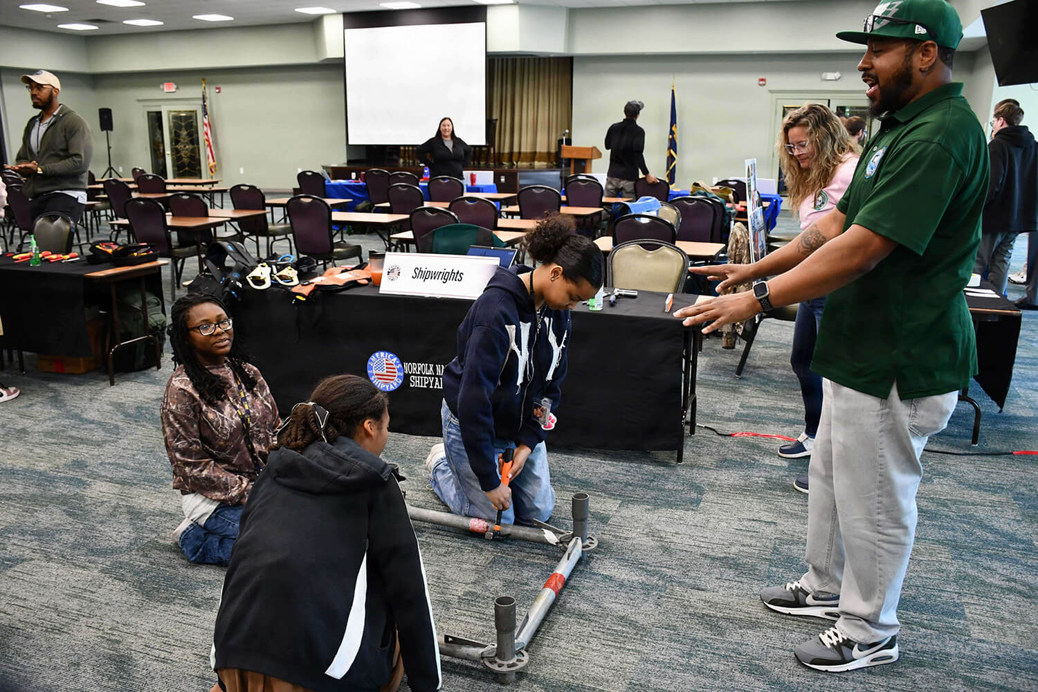 Teens try hands-on shipwright tools at a Norfolk Naval Shipyard apprenticeship event as an instructor guides them.