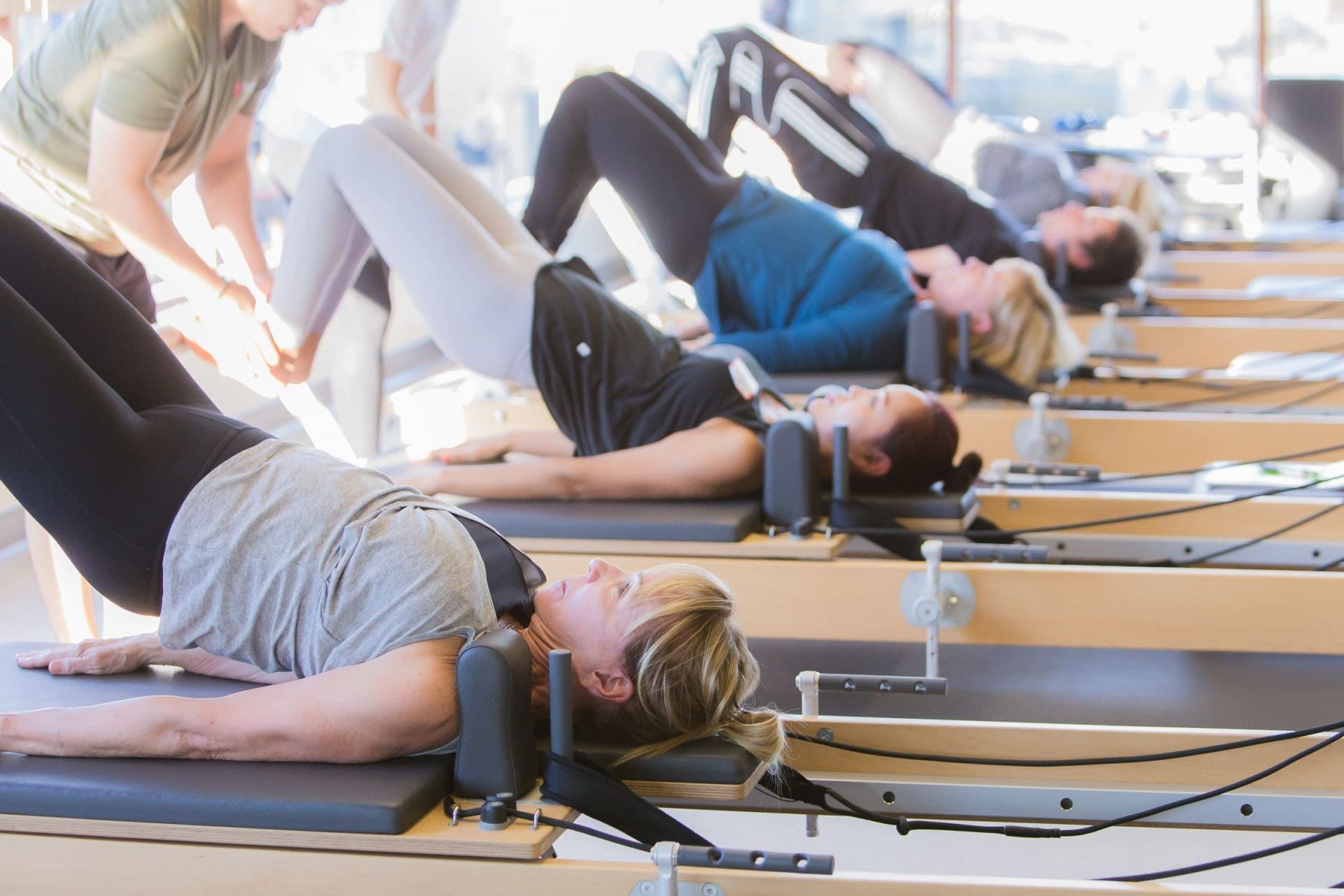 Group of people doing Pilates exercises on reformer machines in a bright fitness studio.