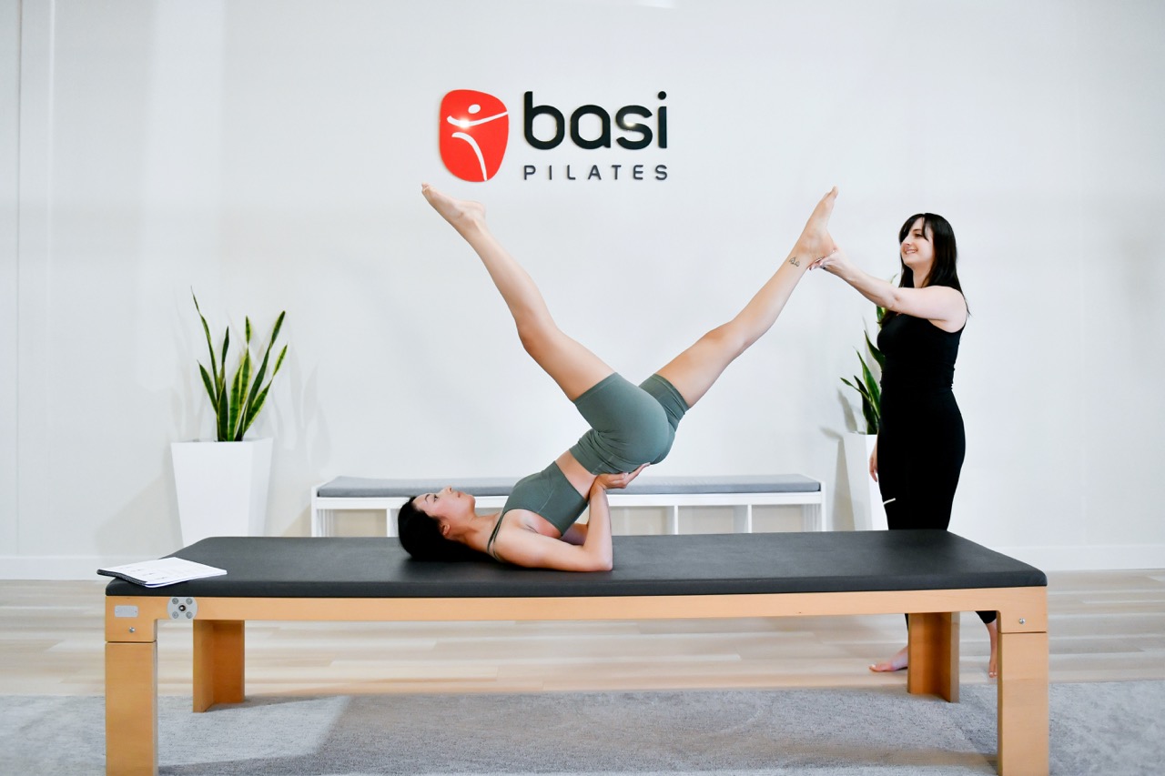 Pilates instructor assists woman performing Scissors mat exercise on a Pilates mat table.