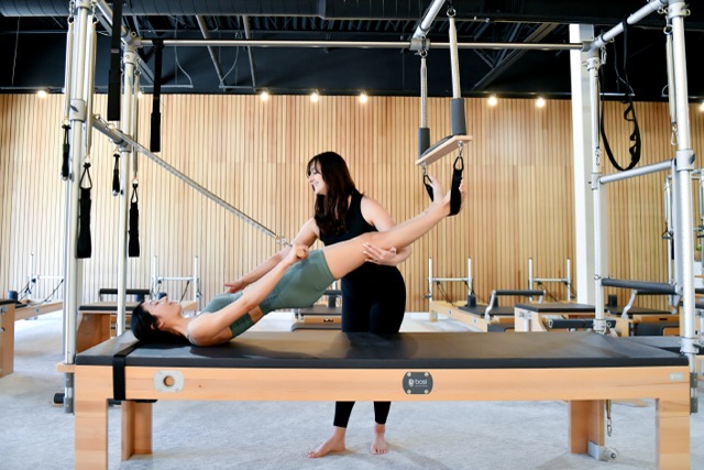 Instructor assisting woman performing Pilates exercise on Cadillac machine in fitness studio.