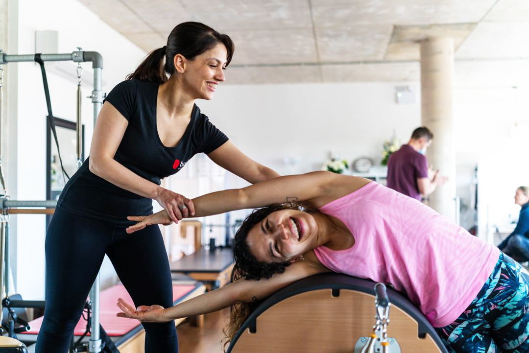 Woman in black guiding another woman in pink doing a stretch on gym equipment, both smiling.