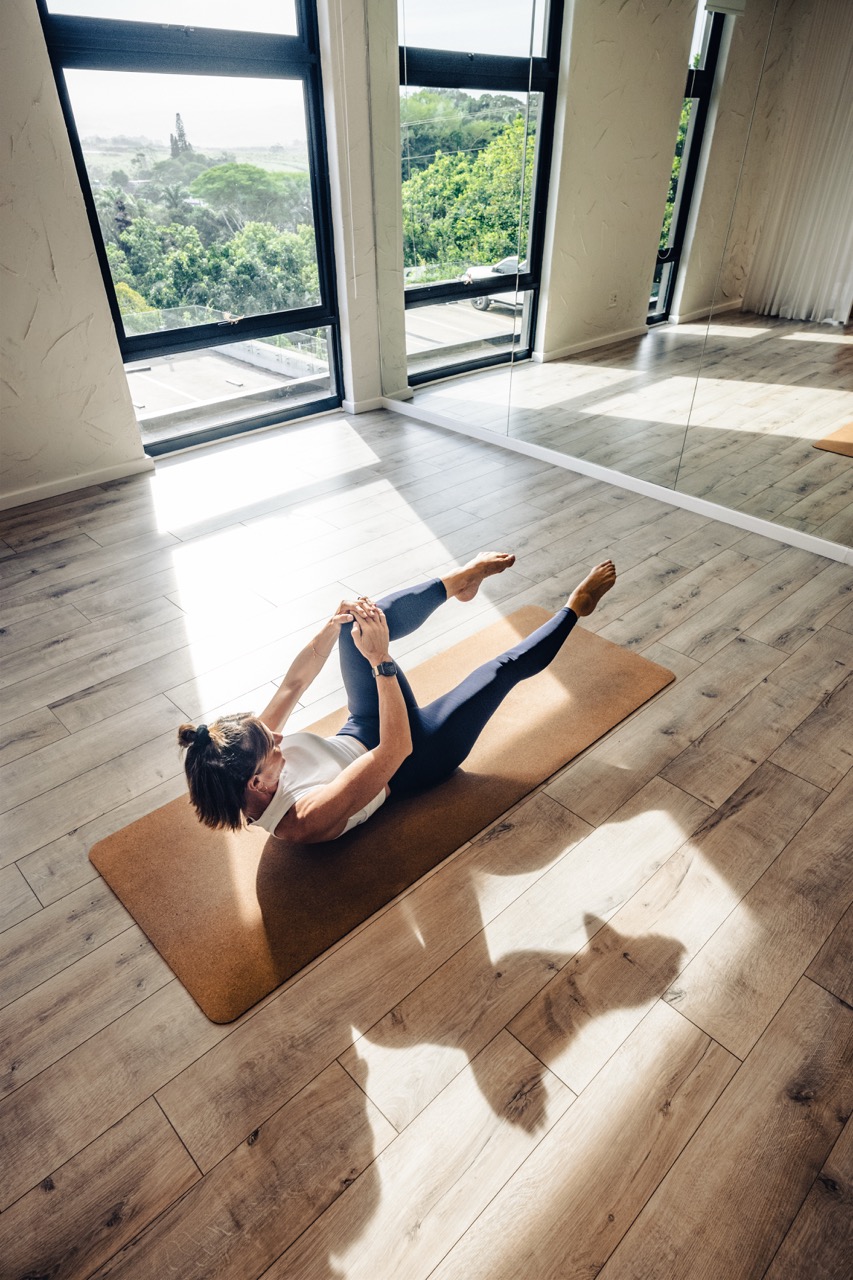 Woman doing Single Leg Stretch Mat exercise on a mat in a sunlit room with large windows and wooden floor.