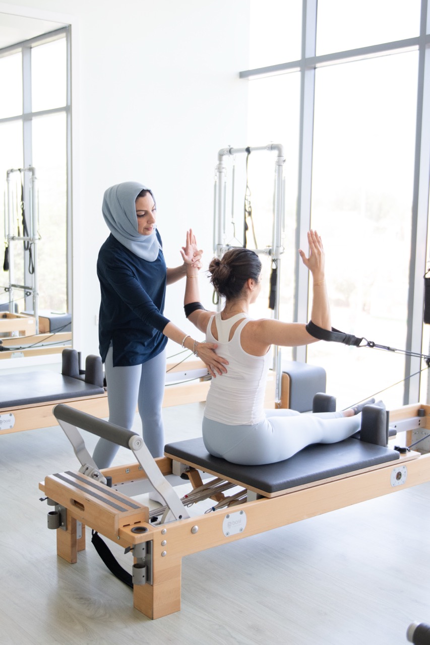 Instructor assists a woman using Pilates reformer machine for arm and back exercise.