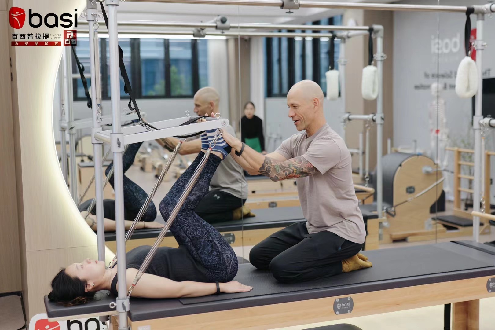 Image of a male Pilates instructor smiling as he helps a student perform an exercise on the Pilates Cadillac.