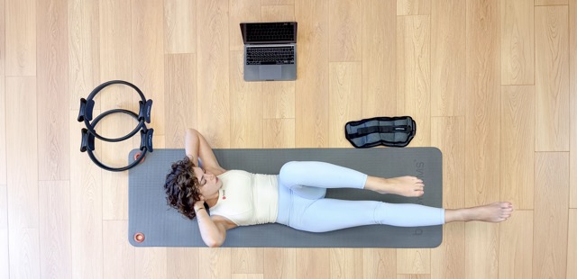Woman lying on a gray mat doing Chest Lift with Rotation Mat exercise near a laptop and Pilates rings on wooden floor.