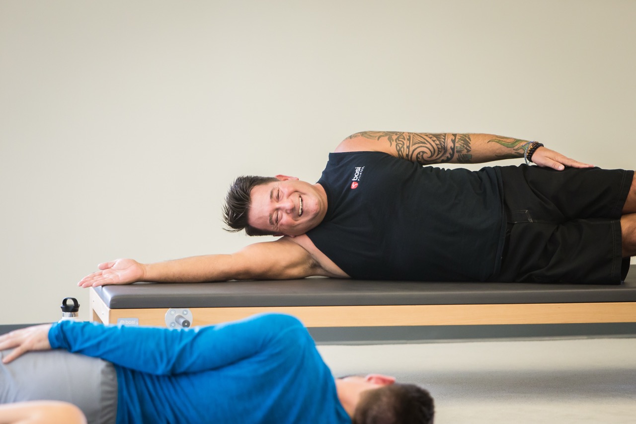 Man with tattoos lying sideways on a Mat table stretching arm, another person in blue beside him.