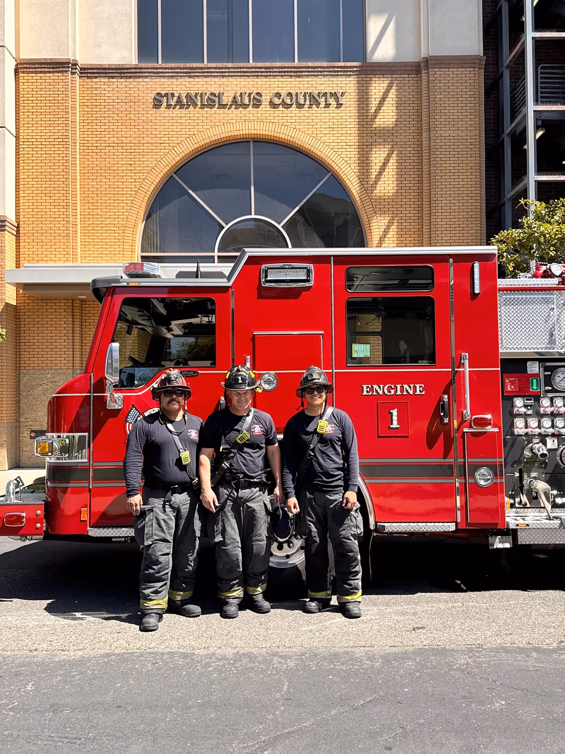 Three firefighters in gear standing in front of a red fire engine truck at Stanislaus County building.