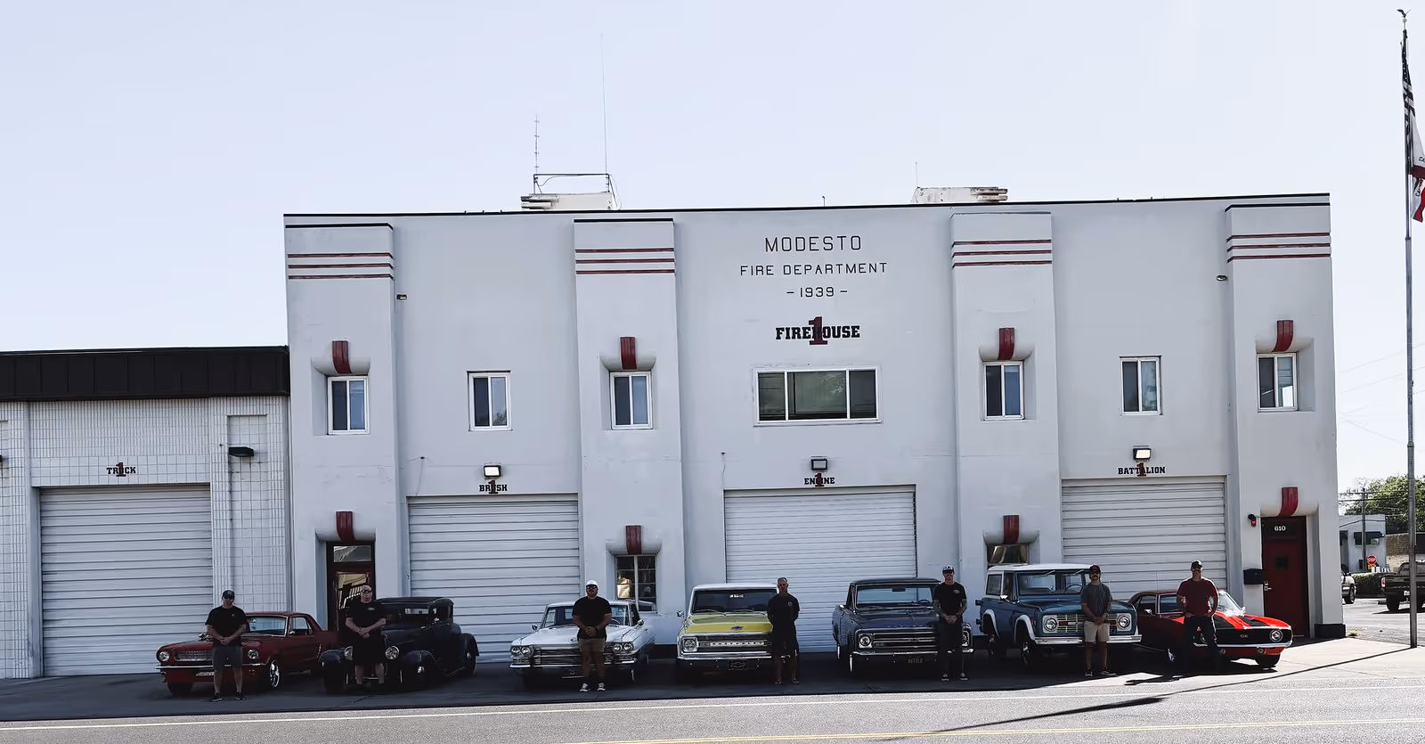 Seven men standing beside vintage cars in front of Modesto Fire Department Firehouse 1 building.