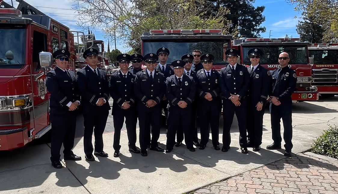 Group of firefighters in uniform standing in front of red fire trucks outdoors on a sunny day.