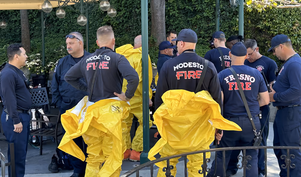 Firefighters from Modesto Fire and Fresno Fire departments stand together outdoors under a pergola, wearing dark uniforms. Several yellow protective hazmat-style suits are draped over a railing in the foreground.