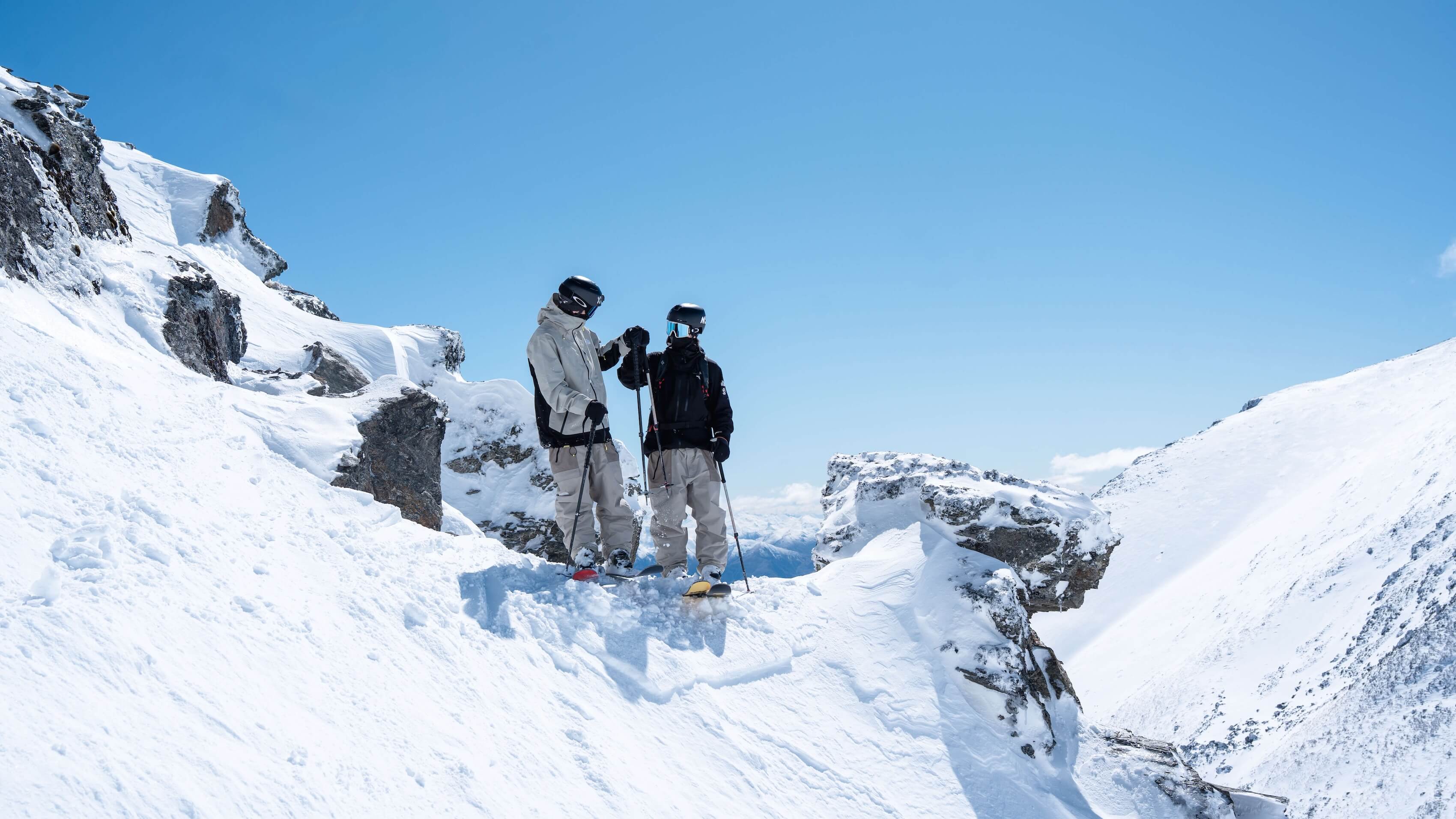 Two skiers fist bump on a snowy mountain ridge under a clear blue sky.