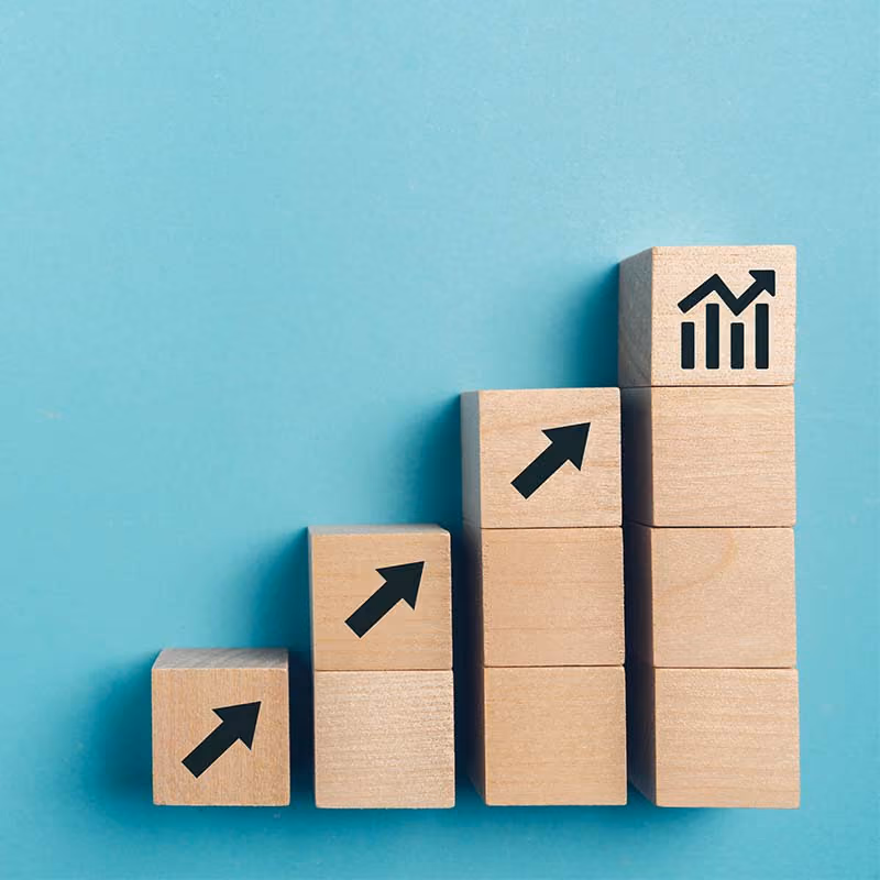 Stacked wooden blocks with upward arrows showing increasing growth, last block has a bar chart with rising arrow.