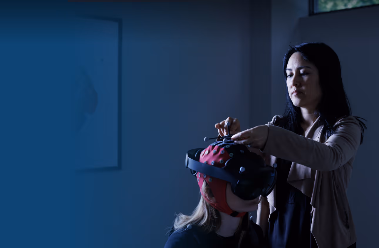 A research participant seated wearing a mobile scalp EEG system and virtual reality headset. Dr. Suthana assisting the participant in putting on the equipment.