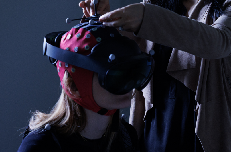 A research participant seated wearing a mobile scalp EEG System and virtual reality headset. A pair of hands assisting the participant in putting on the equipment.