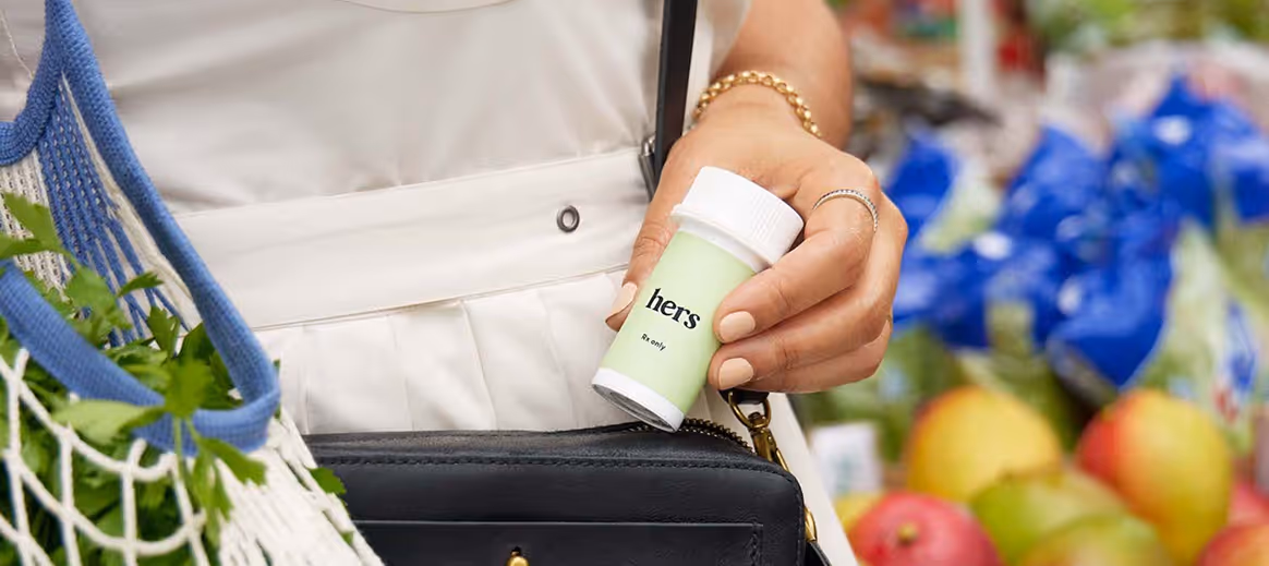 Person holding a green prescription bottle labeled Hers near a black purse and a mesh bag with greens.