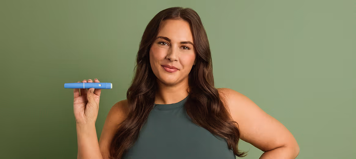 Woman with long brown hair in a green top holding a GLP-1 medication pen against a green background.