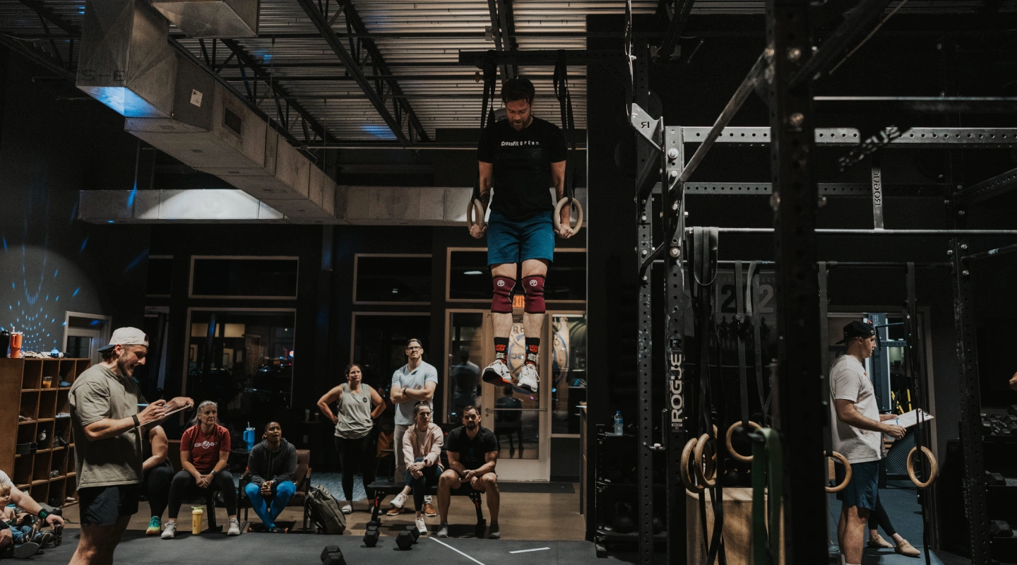 A person is performing ring dips in a gym, surrounded by spectators. The gym is dimly lit, creating an intense and focused atmosphere.