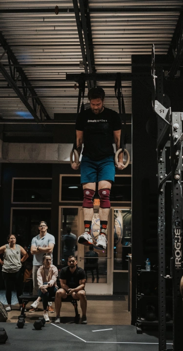 A person is performing ring dips in a gym, surrounded by spectators. The gym is dimly lit, creating an intense and focused atmosphere.