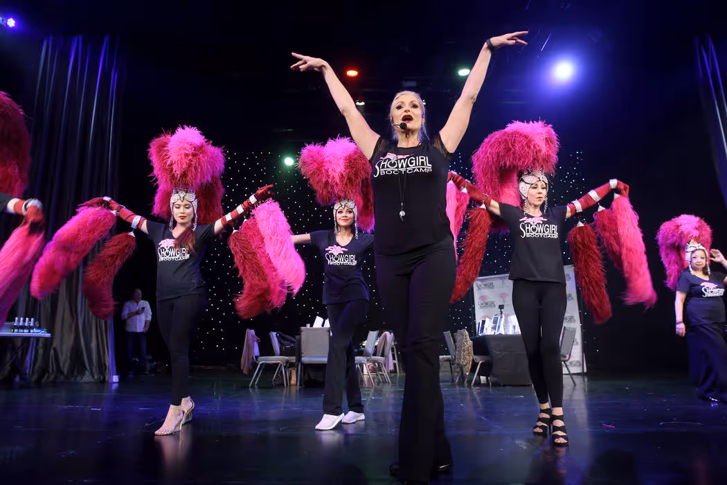 Women wearing Showgirl Bootcamp shirts and large pink feather headpieces dancing on stage.