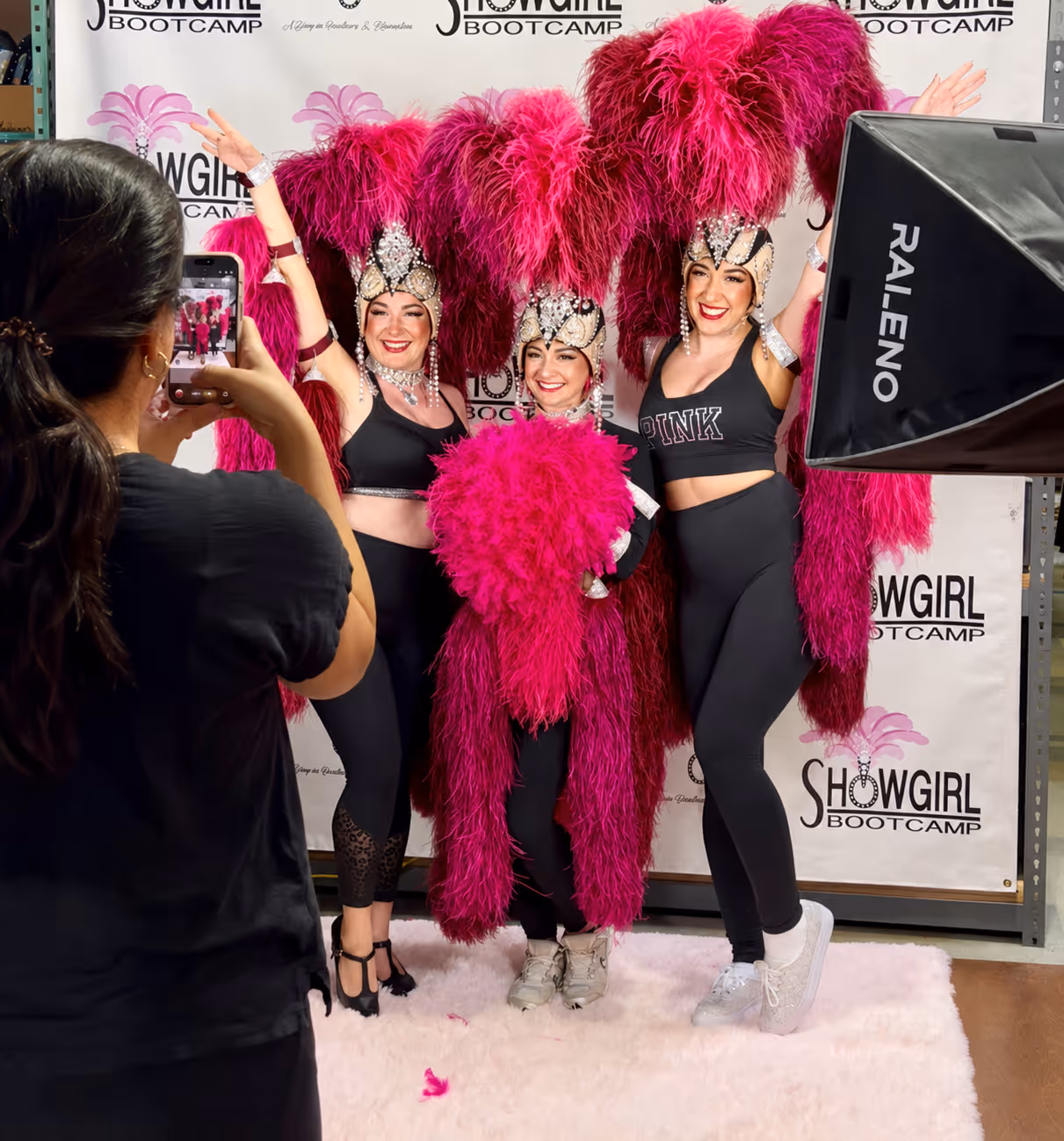 Three women in black outfits and pink feathered headpieces pose for a photo at Showgirl Bootcamp in Las Vegas Nevada.