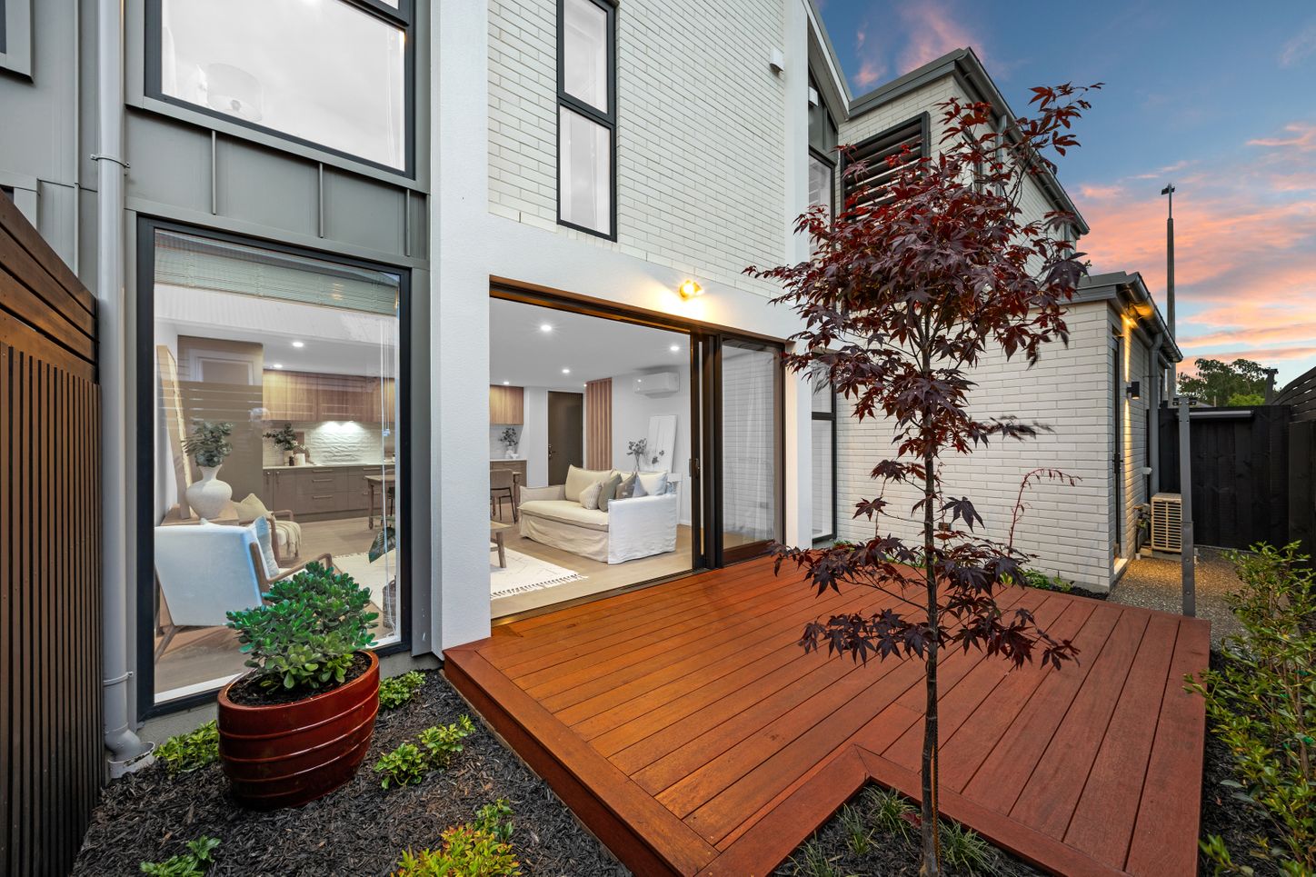 Modern house with sliding glass doors opening to a wooden deck, small tree, and potted plant.