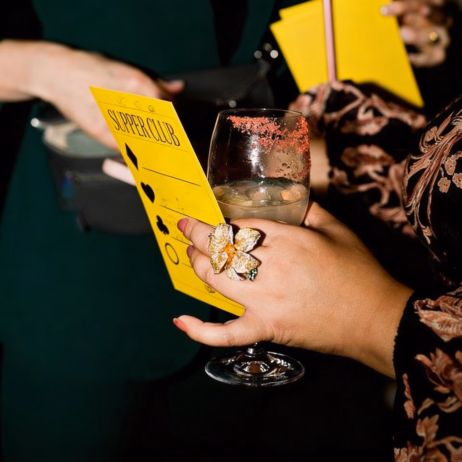 image generated by magnific AI  - Hand with a large flower ring holding a glass with pink salt rim and a yellow Supper Club card.