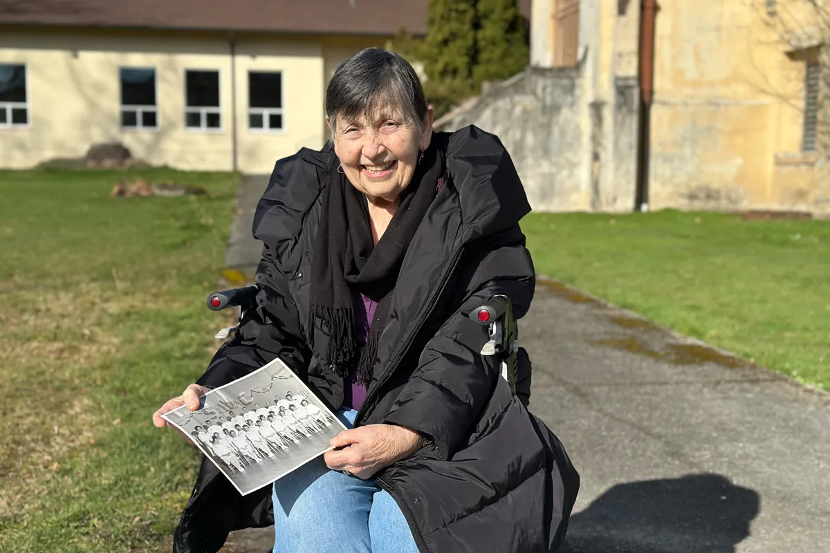 Smiling elderly woman in black coat seated outdoors holding a black-and-white group photo.
