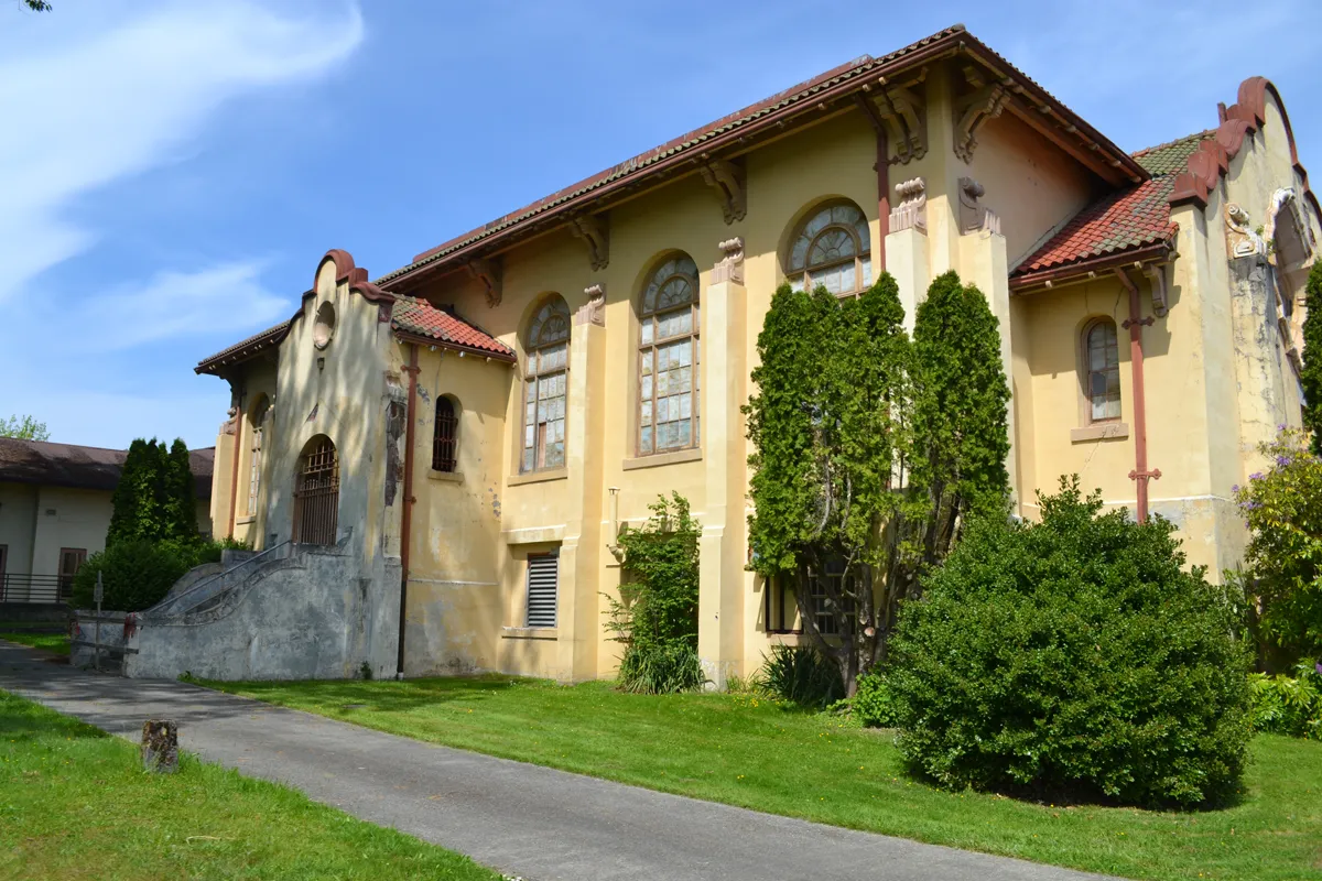 Yellow stucco building with arched windows and red tile roof surrounded by green lawn and bushes.