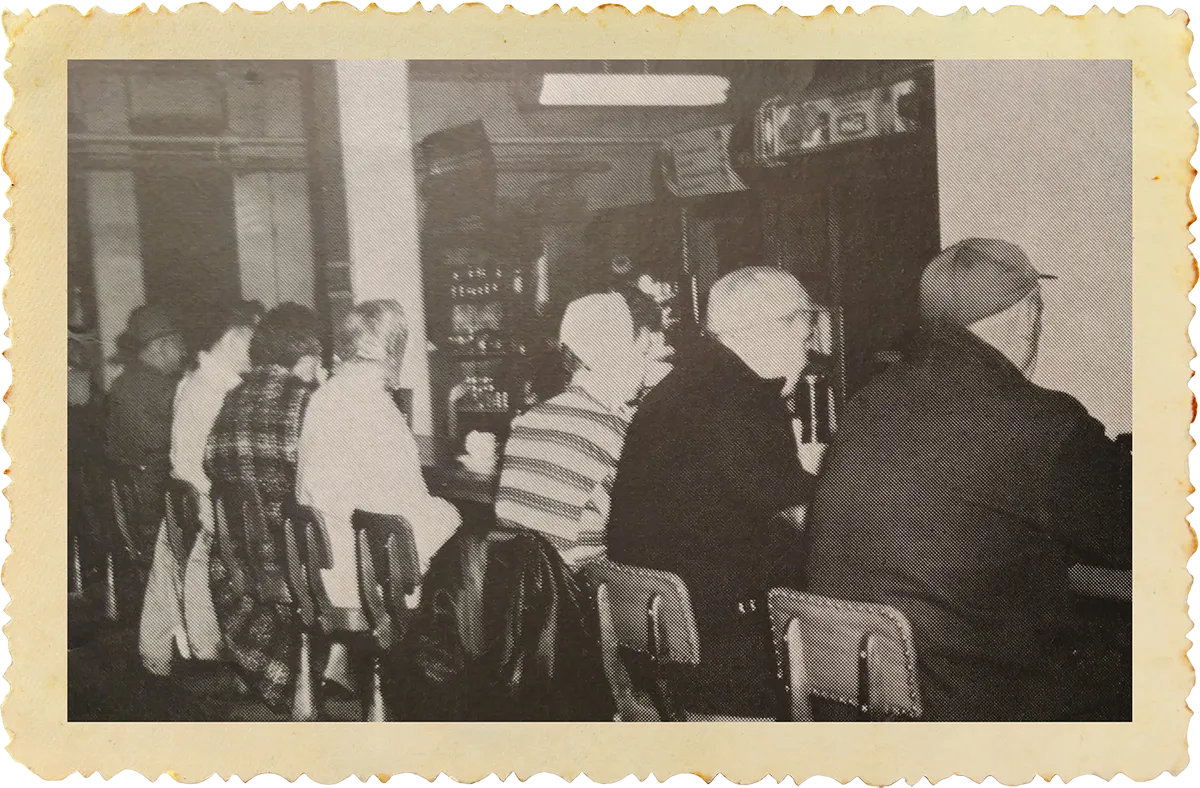 Black and white photo of people sitting side-by-side at a bar or counter on wooden chairs.