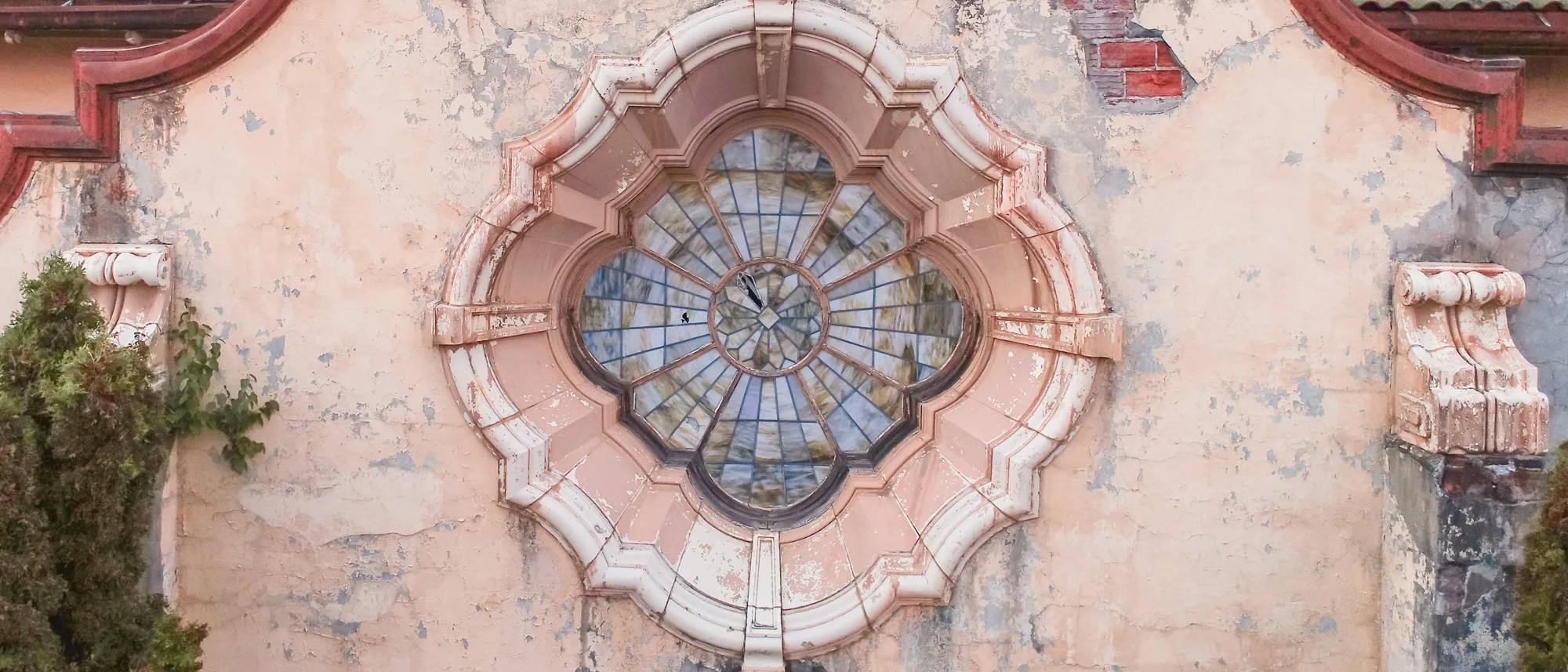 Ornate round stained glass window on aged, peeling peach wall with greenery on sides.
