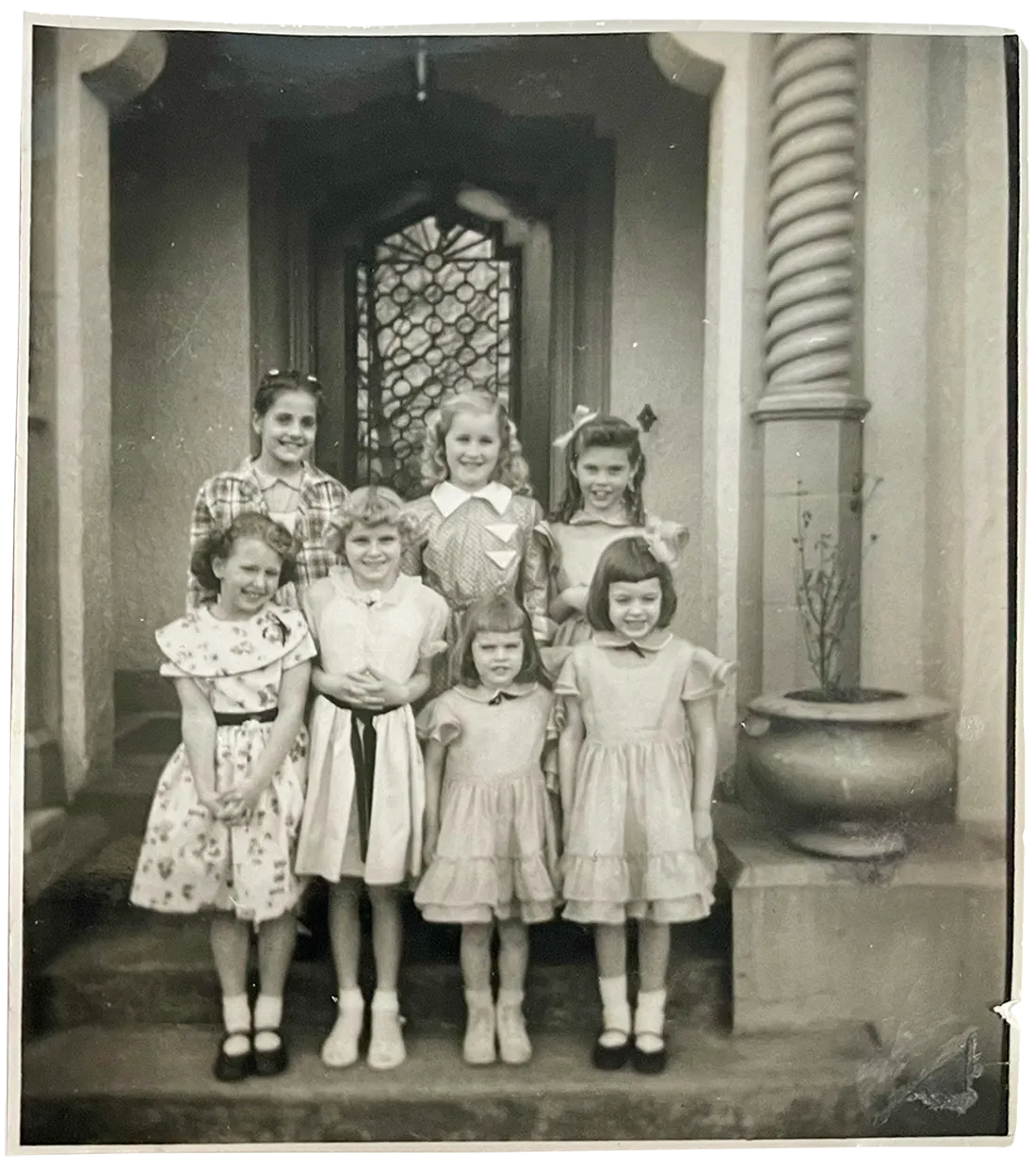 Seven young girls in dresses posed on steps outside a building with decorative door and columns.
