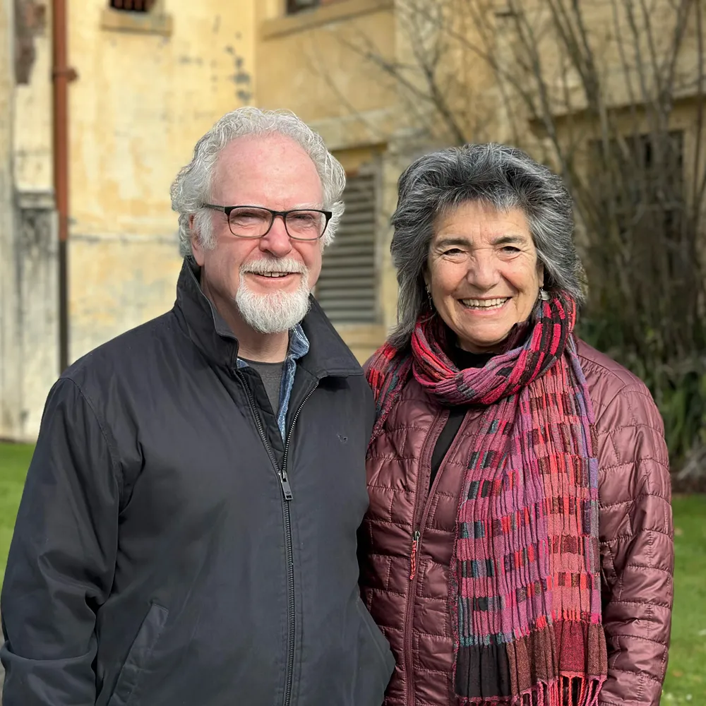 Smiling elderly man with glasses and woman in colorful scarf stand outdoors in front of a beige building.